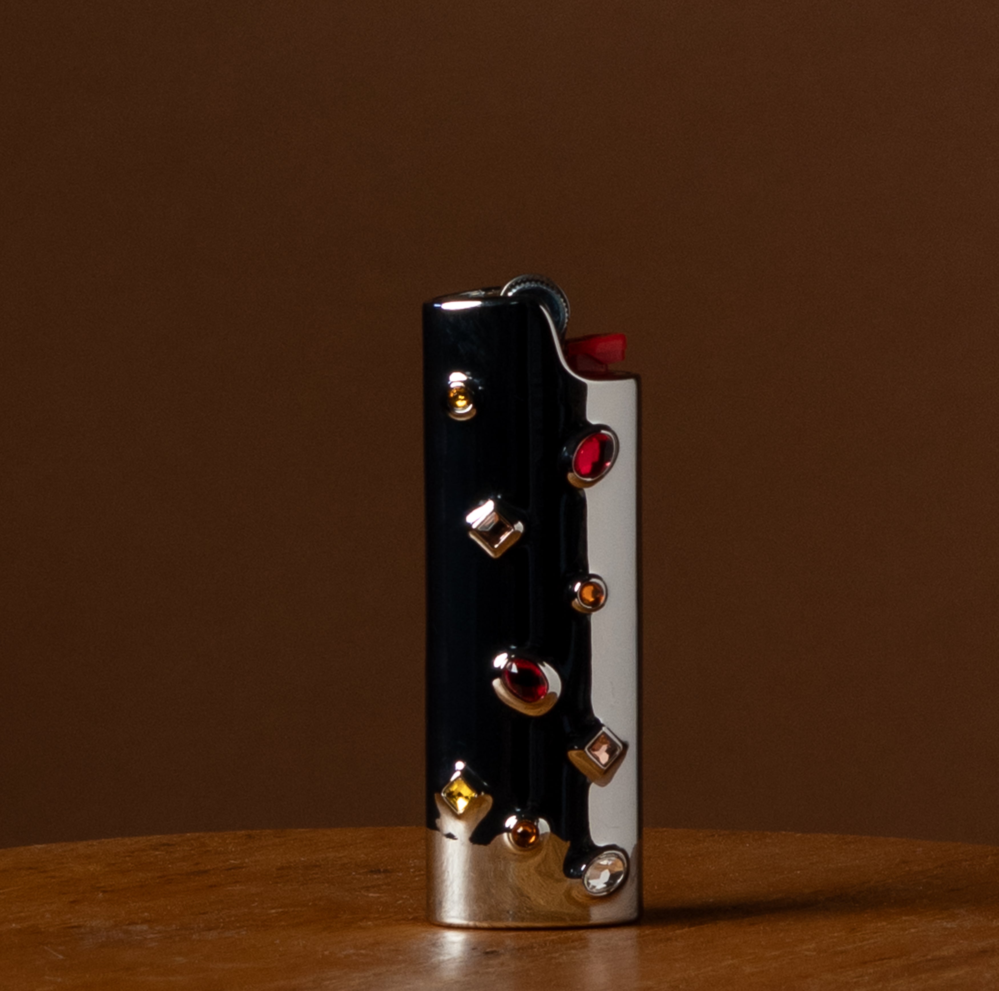 A luxury lighter with a black and silver body, embellished with multiple red and gold gemstones, standing on a wooden surface against a solid brown background.